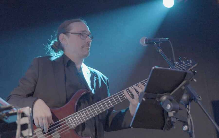 Ein Musiker mit Brille und im dunklen Anzug spielt Bassgitarre auf einer Bühne. Er steht neben einem Mikrofon und einem Notenständer mit einem Tablet. Die Atmosphäre ist von sanftem, blauem Bühnenlicht geprägt, welches von LemonPics eingefangen wurde.