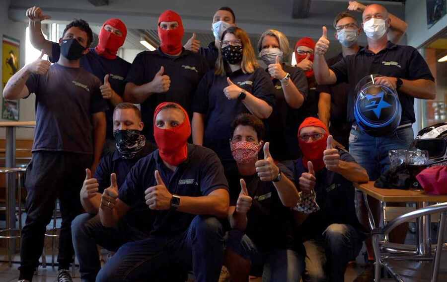 Gruppe von Personen in einem Raum, alle tragen verschiedene Gesichtsmasken und geben mit ihren Daumen ein positives Zeichen. Einige tragen rote Sturmhauben, während andere chirurgische Masken oder Stoffmasken tragen. Ein Mann hält einen Motorradhelm mit einem Blitz-Symbol.