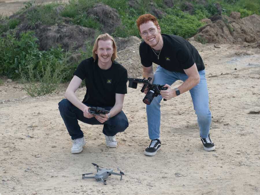 Zwei Männer im Freien, beide tragen schwarze 'LemonPics' T-Shirts. Einer hält eine Kamera, während der andere eine Drohnenfernbedienung hält. Im Hintergrund sieht man ein trockenes Gelände mit Büschen.