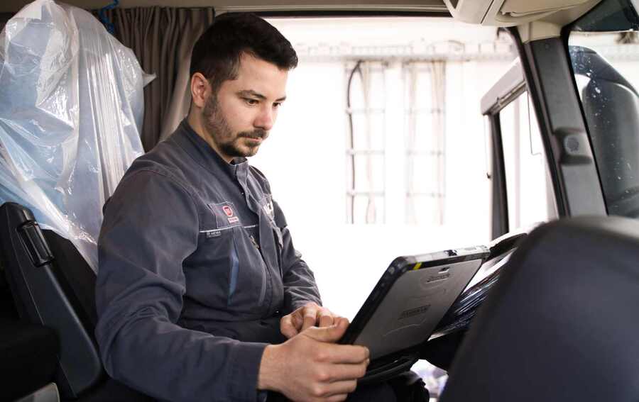 "Ein IVECO-Mitarbeiter in Arbeitsuniform sitzt konzentriert im Fahrerhaus eines Lkw und nutzt ein Tablet zur Überprüfung oder Einstellung. Das Bild unterstreicht die moderne Technik und den Fachkompetenz des Personals.