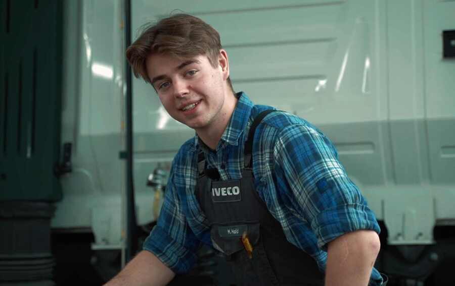 Ein junger IVECO-Mitarbeiter  lächelt freundlich in die Kamera. Er trägt ein kariertes Hemd und eine Arbeitsweste mit IVECO-Logo. Im Hintergrund ist ein Teil eines Lkw zu sehen. Der Mitarbeiter strahlt eine jugendliche Energie und Professionalität aus.